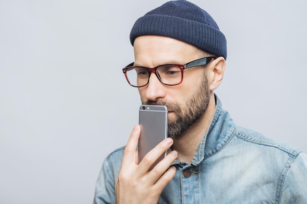 Skupiony mężczyzna w czapce i okularach trzymający smartfon przy uchu zaangażowany w rozmowę z neutralnym szarym tłem