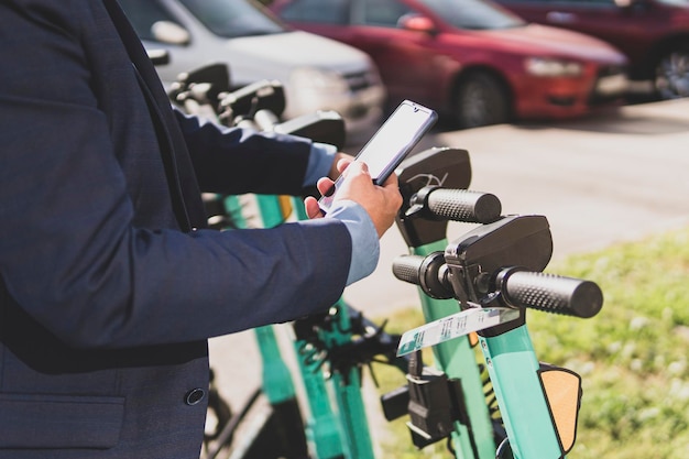 Skuter elektryczny w mieście Mężczyzna używający smartfona do wypożyczenia e-pojazdu Facet z telefonem komórkowym korzystający z ekologicznego transportu w centrum Biznesmen w ruchu ulicznym Nowoczesny miejski styl życia latem