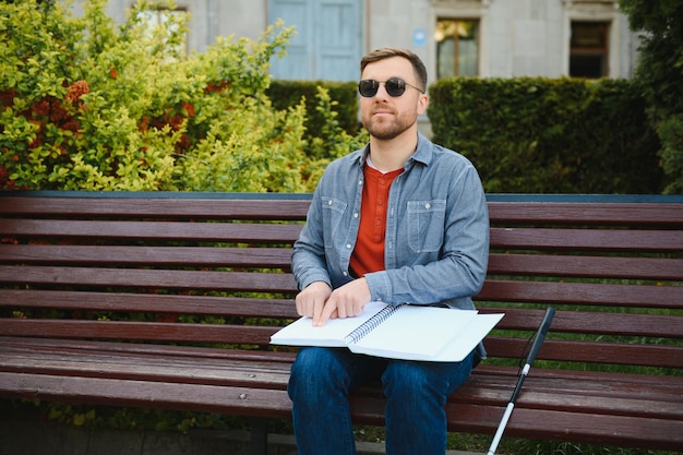 Ślepiec czytający książkę Braille'a, siedzący na ławce w letnim parku, odpoczywający