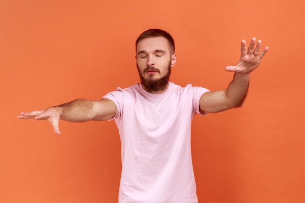Zdjęcie Ślepota zagubiona w ciemności mężczyzna z zamkniętymi oczami i wyciągniętymi rękami szuka drogi, czując się sfrustrowany i zdezorientowany, ubrany w różową koszulkę indoor studio shot na białym tle na pomarańczowym tle