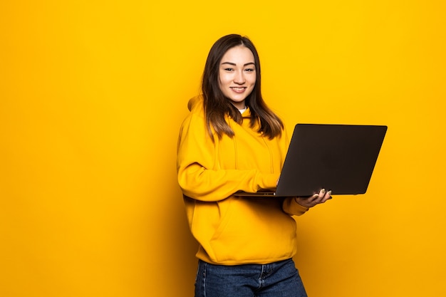 Śliczna azjatycka kobieta studiująca na laptopie i uśmiechnięta, stojąca przy żółtej ścianie