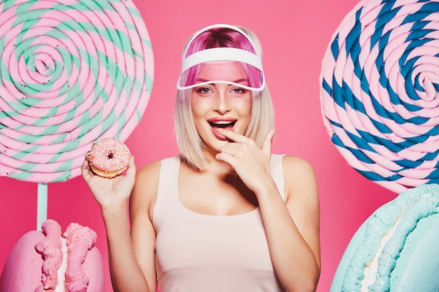 Śliczna dziewczyna o blond włosach, ubrana w bluzkę i różową czapkę, stojąca z ogromnymi słodkimi lizakami w różowym studio
