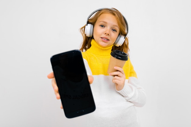 Śliczna dziewczyna pokazuje ekran smartfona z filiżanką kawy w jej rękach i słuchawkami na głowie