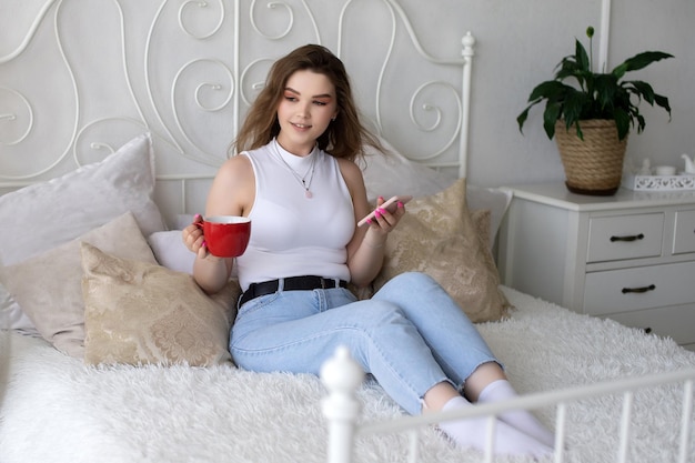 Śliczna dziewczyna siedzi na białym łóżku z telefonem i czerwonym kubkiem
