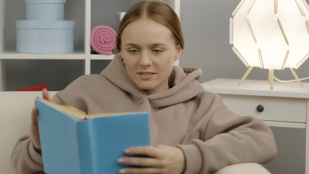 Śliczna dziewczyna siedzi na kanapie i czyta fascynującą książkę edukacyjną podczas kwarantanny