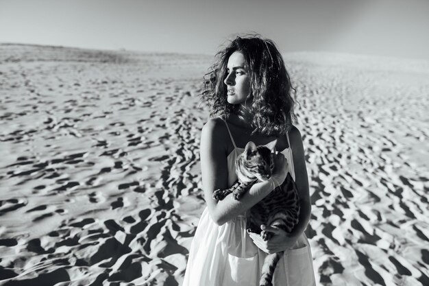 Śliczna dziewczyna w białej sukni na plaży przytula swojego bengalskiego kota