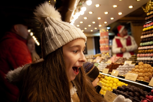 Śliczna dziewczyna w pobliżu słodkiego stoiska z czekoladowymi cukierkami na jarmarku bożonarodzeniowym Święta zimowe obchody tradycji i dzieciństwa