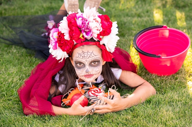 Śliczna dziewczyna z pomalowaną twarzą trzymającą stos słodkich halloweenowych smakołyków, leżąc na zielonym trawniku przed kamerą i relaksując się w słoneczny dzień