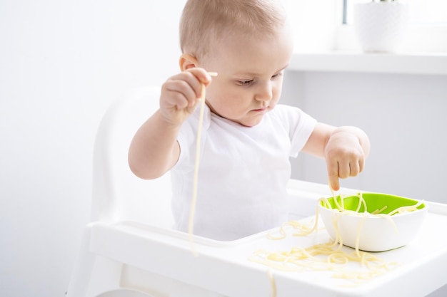 śliczna dziewczynka je makaron spaghetti siedzi w krzesełku dla dziecka na białej kuchni