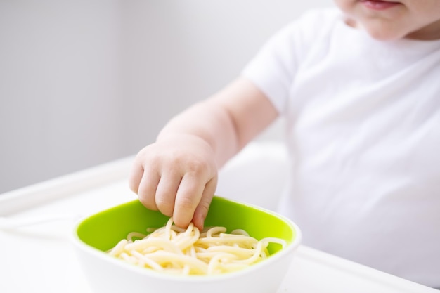 śliczna dziewczynka je makaron spaghetti siedzi w krzesełku dla dziecka na białej kuchni