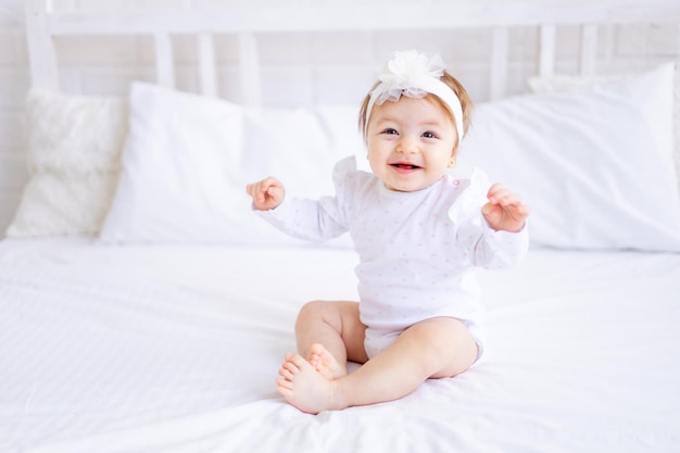 Śliczna dziewczynka siedzi na łóżku w białych ubraniach i z kokardą na głowie śmieszne małe dziecko na bawełnianym łóżku w domu i uśmiechnięta koncepcja produktów dla dzieci