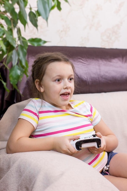 Śliczna dziewczynka trzymająca konsolę do gier i grająca w gry wideo na kanapie w domu