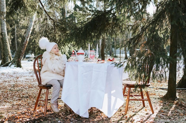 Śliczna dziewczynka w pięknych zimowych ubraniach siedzi przy stole przykrytym białym obrusem na śniadanie na zewnątrz zimą herbata rogaliki i ciasta romantyczna randka na pikniku