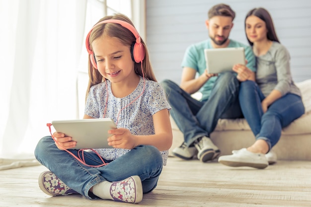 Śliczna dziewczynka w słuchawkach używa tabletu i uśmiecha się, w tle jej rodzice też używają tabletu, siedząc na kanapie w domu