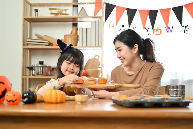 Śliczna i radosna mała Azjatka w fantazyjnym kostiumie robi babeczki na Halloween ze swoją mamą