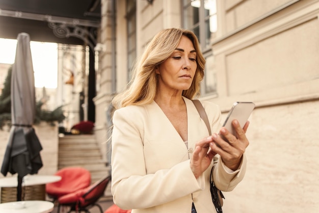Śliczna kaukaska dama w średnim wieku przegląda pocztę na telefonie komórkowym, stojąc na zewnątrz Koncepcja uzależnienia od mediów społecznościowych