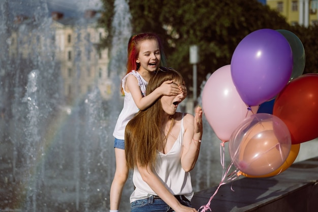 śliczna kaukaska uśmiechnięta dziewczyna z kolorowymi farbowanymi włosami stojąca za mamą zamykającą oczy trzymająca bal