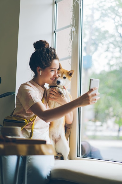 Śliczna kobieta biorąca selfie z psem shiba inu siedzącym na parapecie w kawiarni