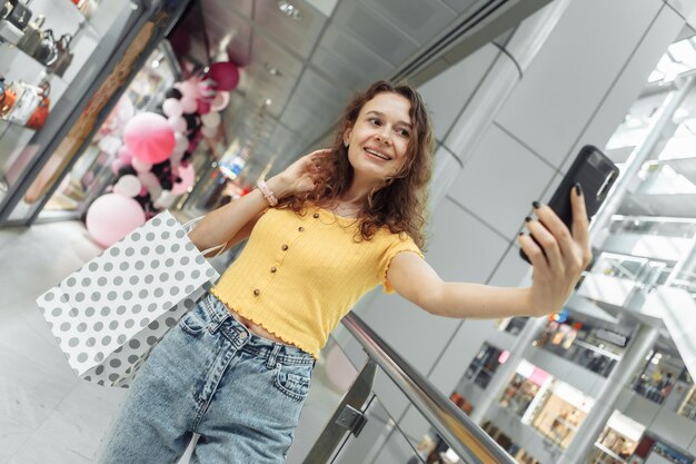 Śliczna kobieta z kręconymi włosami robi selfie z torbami na zakupy w centrum handlowym Koncepcja zakupoholiczki