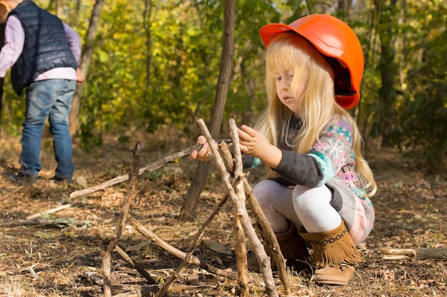 Śliczna mała blond dziewczyna w ogromnym kasku, kucając, konstruując wigwam z naturalnych patyków w lesie