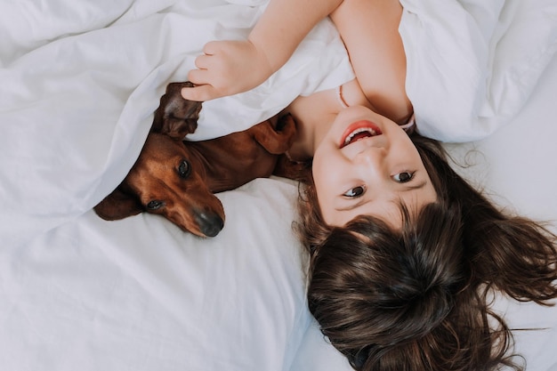 śliczna mała brunetka w domu w łóżku z brązowym jamnikiem przytulającym i śpiącym