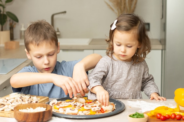 Śliczna mała dziewczynka 2-4 w szarej sukience i chłopiec 7-10 w koszulce gotują razem pizzę w kuchni. Brat i siostra gotują