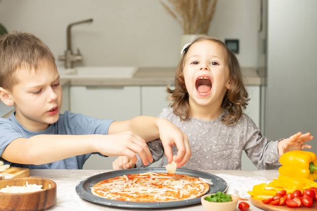 Śliczna mała dziewczynka 2-4 w szarej sukience i chłopiec 7-10 w koszulce gotują razem pizzę w kuchni
