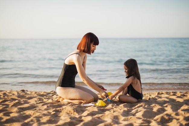 Śliczna mała dziewczynka bawi się z matką siedząc nad brzegiem morza na tropikalnej plaży