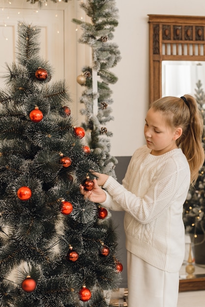 Śliczna mała dziewczynka dekoruje choinki z zabawek czerwonymi piłkami. Dziewczyna w białej dzianinowej sukience swetrowej trzyma wisi piłki na sztucznym świerku