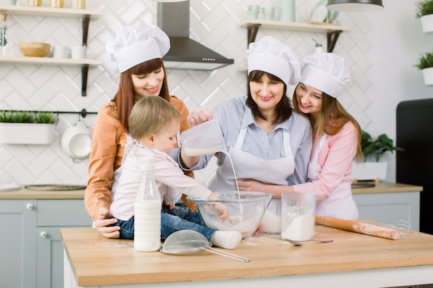 Śliczna mała dziewczynka i jej piękna mama, ciocia i babcia w fartuchach i czapkach bawiących się Podczas nalewania mleka do mąki i ugniatania ciasta w nowoczesnej kuchni w Sweet Home. Kobiety do pieczenia w kuchni