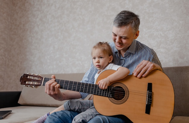 Śliczna mała dziewczynka i jej przystojny ojciec grają na gitarze i uśmiechają się, siedząc na kanapie w domu. dzień Ojca. opieka i edukacja dzieci.