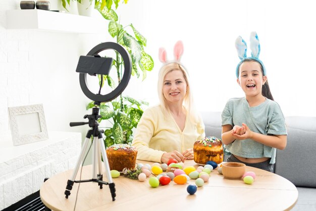 Śliczna mała dziewczynka i piękna młoda mama siedzą razem przy stole w kuchni przy użyciu inteligentnego telefonu online na Wielkanoc.