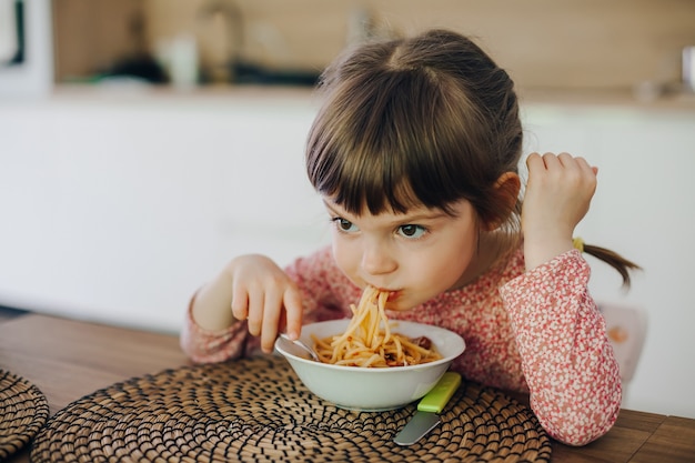 Śliczna mała dziewczynka je spaghetti z sosem, siedząc w kuchni w domu