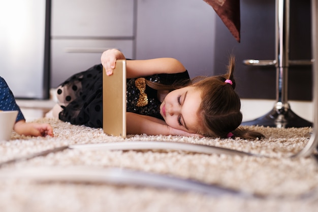Zdjęcie Śliczna mała dziewczynka patrzeje pastylkę z ponytails podczas gdy kłamający na dywanie.