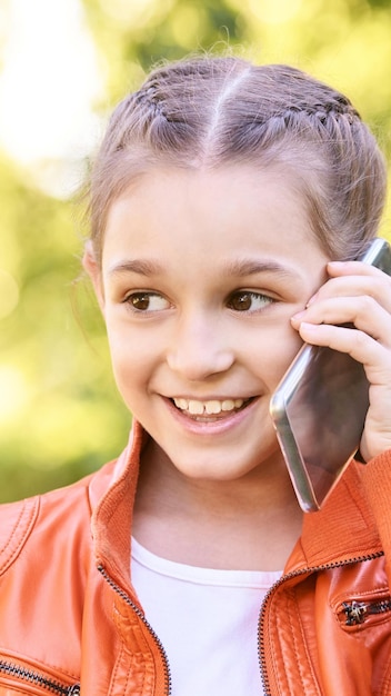 Śliczna mała dziewczynka rozmawia Trzymaj telefon blisko twarzy Uśmiech młodej kobiety Nie patrz na kamerę Szkoła na świeżym powietrzu Zielone tło Uśmiechnięta mowa Szczęśliwy wyraz Studencki styl życia Pionowo