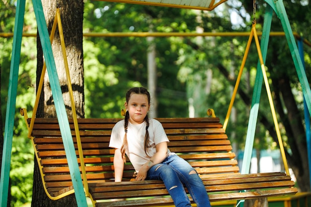Śliczna mała dziewczynka siedzi na drewnianej huśtawce na placu zabaw w zielonym parku Letnie wakacje w centrum turystycznym obozu Spacery i zabawy na świeżym powietrzu, aktywność sportowa i rekreacja zdrowego stylu życia