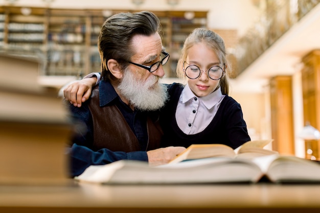 Śliczna mała dziewczynka siedzi przy stołem w antycznej bibliotece w eyeglasses, ściska jej dziadu