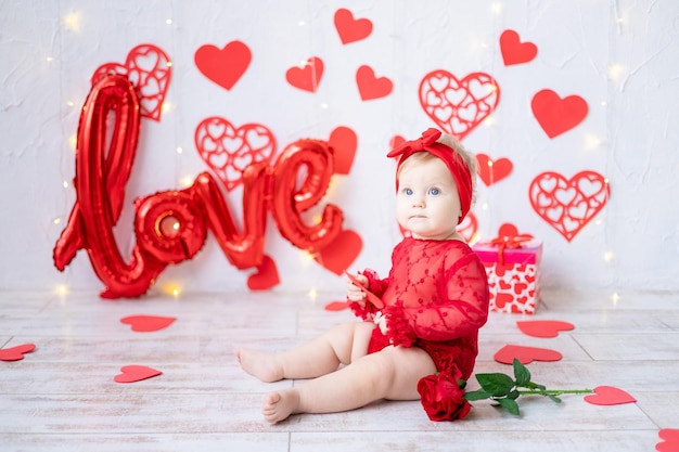 Śliczna mała dziewczynka siedzi w czerwonym body na tle czerwonych serc i napis love koncepcja walentynki walentynki