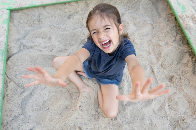 Śliczna mała dziewczynka siedzi w piaskownicy i bawi się piaskiem w upalny letni dzień.