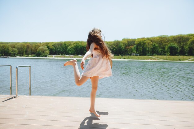 Śliczna mała dziewczynka stoi na molo i patrzy na piękne jezioro, dziecko skacze i bawi się na plaży