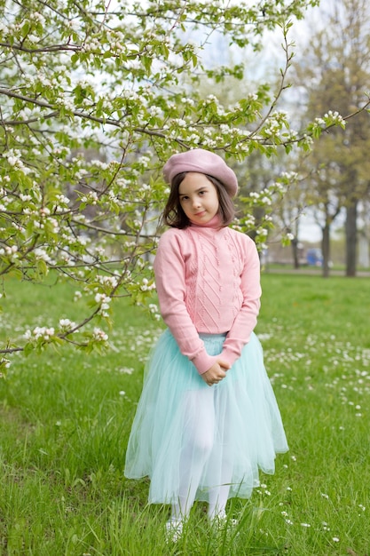 Śliczna mała dziewczynka stoi na zielonej trawie obok kwitnącej białej jabłoni w parku