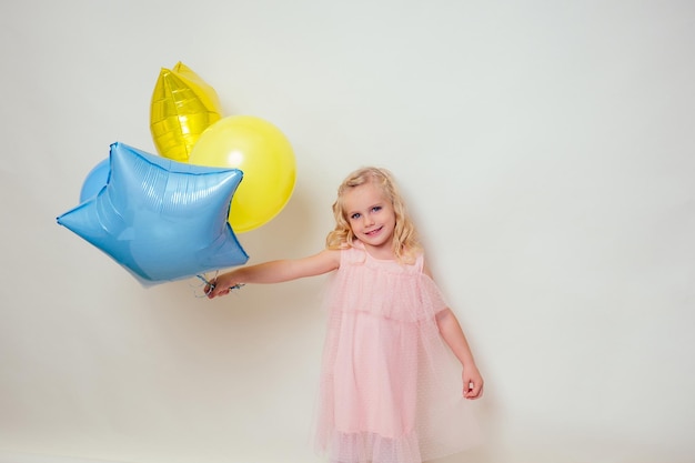 Śliczna mała dziewczynka trzyma helowe niebieskie, żółte i złote balony na białym tle w studio