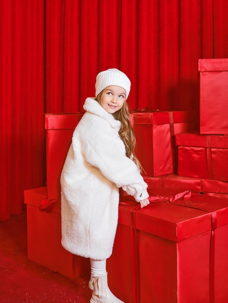 Śliczna mała dziewczynka w białym płaszczu przy ogromnych czerwonych pudełkach świątecznych na czerwonym tle. Wakacje