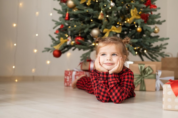 śliczna mała dziewczynka w czerwonej piżamie leżąca na podłodze w pobliżu choinki z pudełkami na prezenty w domu