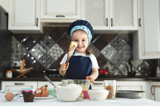 Śliczna mała dziewczynka w fartuchu i czapce szefa kuchni miesza ciasto drewnianą łopatką