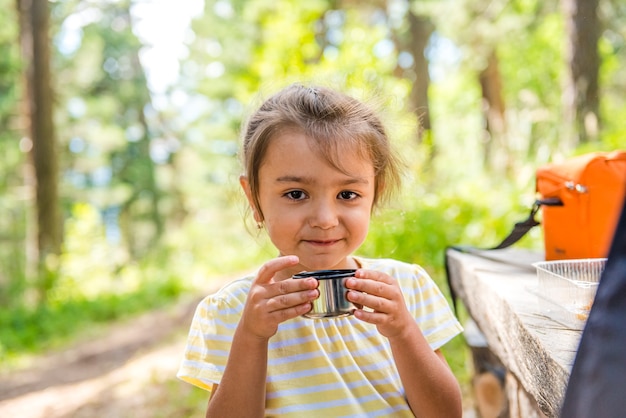 śliczna mała dziewczynka w letnim lesie trzymająca filiżankę herbaty i uśmiechnięta