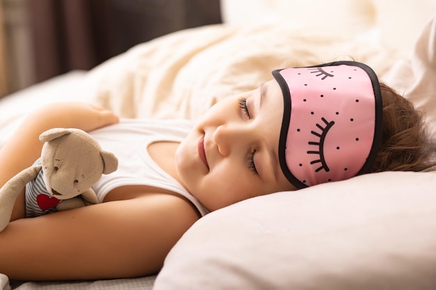 Śliczna mała dziewczynka w nocnej masce leżąca na łóżku i śpiąca z zabawką pluszowego misia