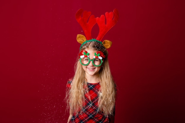 Śliczna mała dziewczynka w okularach Boże Narodzenie wieje śnieg z dłoni w studio na czerwonym tle. Koncepcja Bożego Narodzenia, miejsca na tekst
