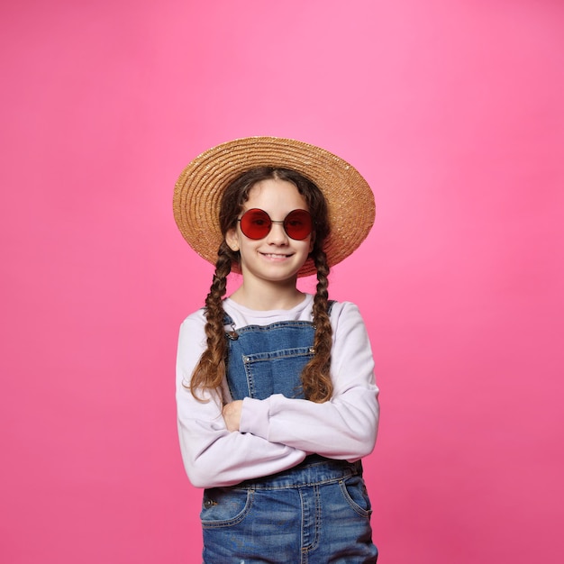 Śliczna mała dziewczynka w słomkowym kapeluszu i czerwonych okularach przeciwsłonecznych patrząca na kamerę ze skrzyżowanymi rękami na białym tle różowego tła