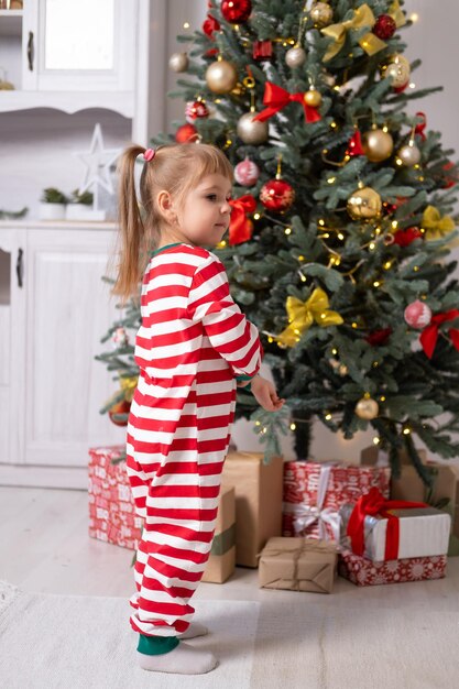 śliczna mała dziewczynka w świątecznej piżamie z pudełkami prezentowymi pod choinką w domu